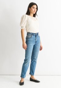 Blusa bianca con maniche a palloncino e pois testurizzati, abbinata a jeans di denim blu a vita alta e ballerine nere, in piedi su uno sfondo bianco.
