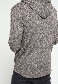 Huppari harmaata sävyä, jossa on teksturoitu kangas, hupullinen malli, pitkät hihat ja rento istuvuus. Hienovarainen kuvio tummista ja vaaleista langoista.