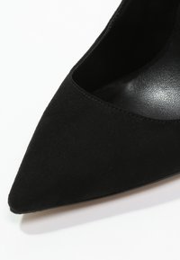 Närbild av en svart mocka spetssko med hög klack och en slät innersula på en vit bakgrund.