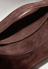 Sac à main en daim marron en gros plan montrant deux rabats qui se chevauchent avec des coutures détaillées et une texture douce sous un éclairage doux.