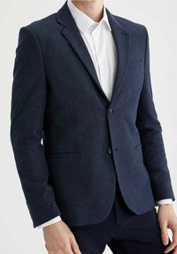 Blazer blu navy in tessuto texture, chiusura a due bottoni, rever a lancia e due tasche laterali. Indossato sopra una camicia bianca, vestibilità su misura.