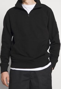 Man in zwart half-zip pullover over een witte shirt met zwarte broek, staande met één hand in zijn zak tegen een effen achtergrond.