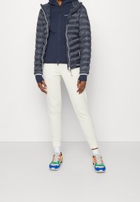 Grau gepufferte Jacke mit Steppdesign, marineblaue Zip-Up-Hoodie, cremefarbene Hose und mehrfarbige Sneakers mit kräftigen Akzenten und geschichteten Texturen.