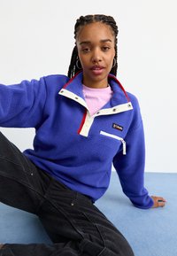 Fleece pulover u živopisnoj plavoj boji s bijelim ovratnikom i crvenim detaljima. Sadrži džep na zip i opušteni kroj. Uparen s crnim trapericama.