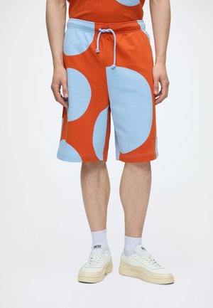 Uomo che indossa pantaloncini arancioni e azzurri a fantasia con coulisse, calzini bianchi e sneakers bianche Diesel, in piedi davanti a uno sfondo neutro.