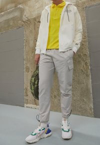 Bílá bunda na zip přes žluté polo, světle šedé kalhoty s boční kapsou a bílé tenisky s barevnými akcenty. Nese zelenou tašku.