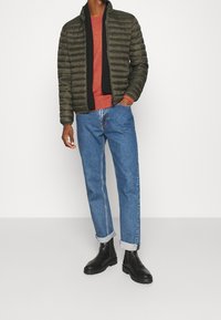 Veste matelassée verte avec col haut, sweat-shirt rouge et jean bleu retroussé. Bottines noires avec semelles texturées. Style minimaliste.