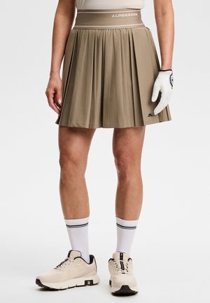 Beige geplooide rok met J.Lindeberg-logo, gedragen met witte golfhandschoen, witte sokken met zwarte streep en beige sportschoenen.