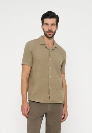 Hombre con camisa beige de manga corta abotonada y pantalones marrones, de pie frente a un fondo blanco liso, mirando hacia adelante.