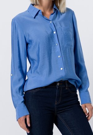 Chemise bleu clair à manches longues avec une finition texturée, une poche poitrine unique et des boutons en ton argenté. Associée à un jean foncé.