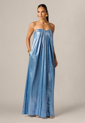 Kobieta w niebieskiej metalicznej sukni bez ramiączek sięgającej do podłogi z plisami, złotymi kolczykami i dopasowanymi bransoletkami, stojąca na jednolitym beżowym tle.