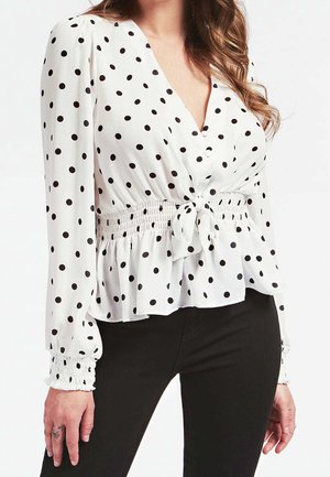 Kvinde iført en hvid bluse med lange ærmer og sorte polkaprikker, V-udskæring, knude foran, rynket talje og sorte bukser.