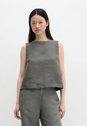 Femme aux longs cheveux noirs portant un haut en lin gris sans manches et un pantalon assorti, debout devant un fond blanc uni.