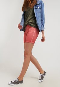 Shorts di jeans in un corallo sbiadito, caratterizzati da un taglio classico e chiusura a bottone frontale, abbinati a una maglietta larga di oliva e sneakers grigie.
