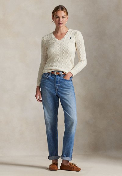 Femme debout portant un pull crème à col en V en maille torsadée, un jean bleu avec des revers, une ceinture marron et des mocassins marron.