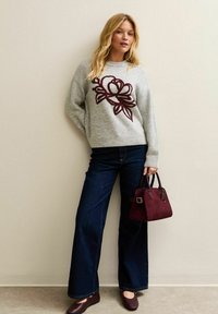 Maglione grigio con ricami floreali rosso scuro, jeans a campana blu scuro, borsa color vino e scarpe marroni scure. Texture liscia e stile casual.