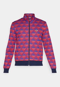 Veste à motifs géométriques rouges et bleus, fermeture éclair, col montant, poignets et ourlet côtelés bleu marine, avec un petit logo vert sur la poitrine.