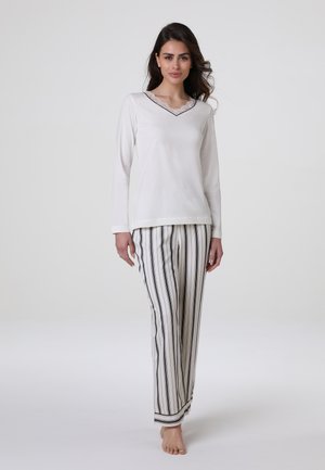 Donna in piedi a piedi nudi, che indossa una maglia bianca a maniche lunghe con bordi in pizzo e pantaloni bianchi con righe nere verticali.