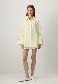 Cămașă oversized de culoare galben deschis, cu guler ascuțit, mâneci lungi cu manșete mari și nasturi rotunzi aurii. Purtată cu pantaloni scurți albi și pantofi.