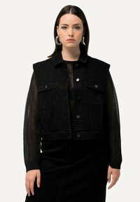 Schwarze Denimweil mit ärmellosem Design, ausgestattet mit zwei Fronttaschen und silbernen Druckknöpfen. Getragen über einem schwarzen transparenten Langarmtop.