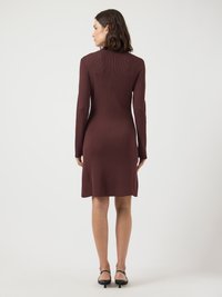 Robe en tricot côtelé bordeaux à manches longues, longueur mi-cuisse et fentes latérales subtiles ; portée avec des escarpins noirs à lanières.