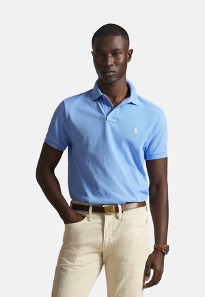 Hombre con polo azul claro con pequeño logo, pantalones beige, cinturón marrón y reloj de pulsera, de pie con la mano en el bolsillo sobre fondo blanco.