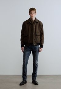 Brun jakke i ullblanding med lommer foran, brukt over en mørk genser, kombinert med mørke jeans og svarte sko, mot en ensfarget bakgrunn.