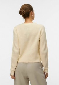 Maglione beige chiaro con un filato testurizzato, dotato di scollo rotondo e maniche lunghe. Indossato con pantaloni di colore neutro.