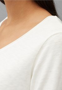 Vit texturerad tyg med en bred V-ringning, mjukt draperad runt nyckelbenen, vilket visar upp ett slätt och lätt material.