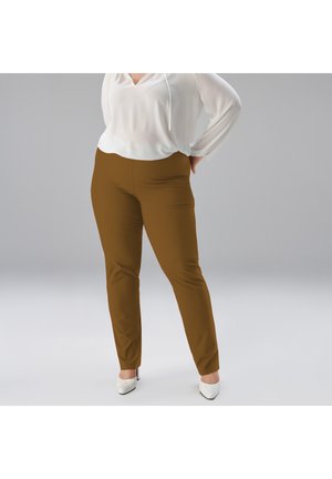 Frau trägt braune Slim-Fit-Hose, weiße Bluse und weiße spitze Absätze, steht mit einer Hand in der Hüfte vor einem einfarbigen Hintergrund.