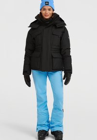 Sort isoleret jakke med flere lommer, parret med lyseblå skibukser. Modellen bærer sorte handsker og en blå beanie.