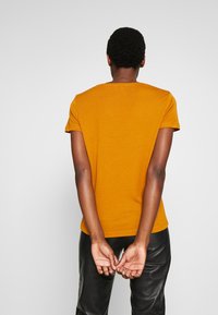 T-shirt a maniche corte color giallo senape con collo tondeggiante, con una texture liscia e una silhouette aderente, mostrato da dietro.