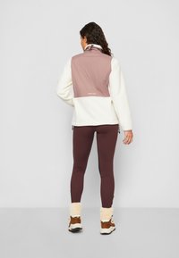 Veste en polaire avec un panneau d'épaule rose clair, corps crème, col ajustable, associée à des leggings bordeaux et des bottes doublées en fourrure marron.