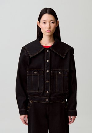 Femme aux cheveux noirs et lisses portant une veste noire à large col et devant boutonné, associée à un pantalon noir assorti, sur un fond uni.