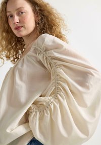 Ljus beige blus med puffärmar, med insamlad detaljering längs sömmarna och en mjuk, slät textur.