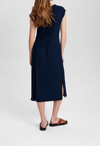 Vestito blu navy al ginocchio con maniche corte, dettaglio arricciato sul retro e spacco laterale. Abbinato a sandali scivolati marroni con suola piatta.