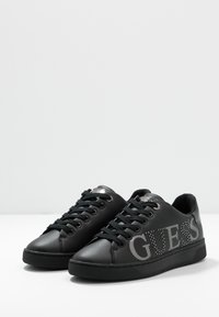 Baskets noires avec une texture lisse en cuir, présentant un logo "GUESS" bien visible et des accents en strass sur le côté, un bout rond et une semelle en caoutchouc.