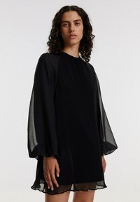 Zwart geplooid mini-jurk met doorschijnende lange mouwen, ronde halslijn en subtiele rimpelzoom. Lichtgewicht, vloeiende stof met een gestructureerde afwerking.