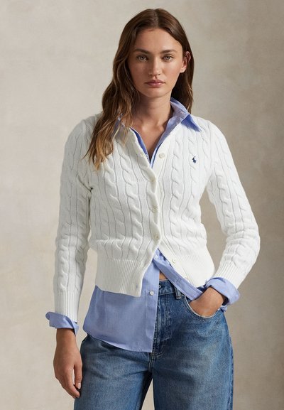 Kvinde stående med den ene hånd i lommen, iført hvid kabelstrikket cardigan over lyseblå skjorte med knapper og blå jeans, neutral baggrund.