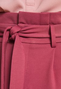 Detailní záběr na uvázaný látkový pásek na kalhotách s vysokým pasem v růžové barvě, se šitým švem a texturovaném růžovém topu nad pasem.