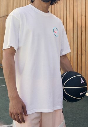 Person hält schwarzen Basketball, trägt weißes Nike-T-Shirt und helle Shorts und steht draußen an einem Holzzaun.