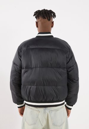 Person mit kurzen Dreadlocks, trägt eine schwarze Steppjacke mit weiß gestreiftem, geripptem Saum und Bündchen sowie hellblaue Jeans, von hinten gesehen.