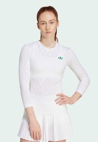 White athletic long-sleeve top with diamond pattern and green logo; paired with a pleated white skirt. Smooth texture, fitted design.