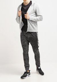 Šedá mikina na zip přes černé tričko spárovaná s texturovanými černými kalhotami s bílými pruhy a černými sportovními botami.