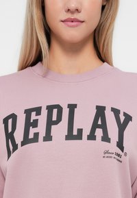 Rožinė megzta palaidinė su rauktiniu aplink kaklą ir juodu "REPLAY" užrašu. Turi minkštą tekstūrą ir mažą logotipo detalę apačioje.