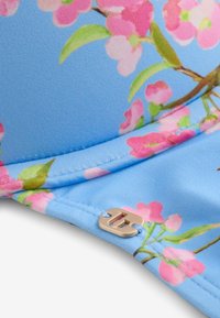 Hellblauer Stoff mit pinkem Blumenmuster und grünen Blättern, versehen mit einem kleinen metallischen Logo, das nahe einer Naht angebracht ist.