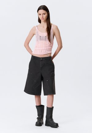 Vrouw met lang donker haar, gekleed in een roze doorzichtige tanktop, zwarte knielange korte broek en zwarte laarzen tot halverwege de kuit, staand met haar handen achter haar rug.