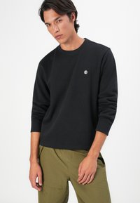 Giovane uomo che indossa una maglietta nera a maniche lunghe con un piccolo logo bianco sul petto e pantaloni verde oliva, in piedi davanti a uno sfondo bianco.