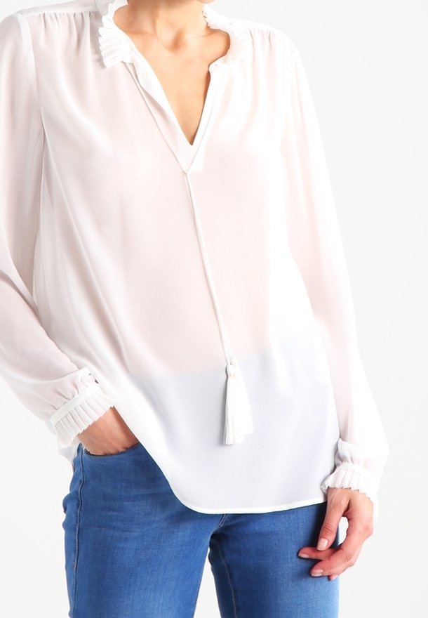 Vit, lätt blus med V-hals, plisserad krage och kvastdetalj. Långa ärmar med samlade manschetter. Matchat med blå jeans.