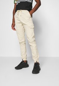 Pantalons cargo beige avec des poignets élastiques, des poches latérales et une taille à cordon, associés à des baskets noires et une chemise noire à manches courtes.